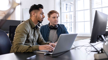 Das Bild zeigt zwei Männer, die in einen Bildschirm schauen, einer von Ihnen hat einen Laptop