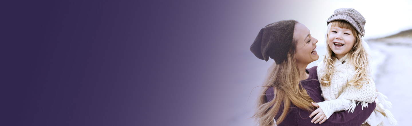 Frau mit langen blonden Haaren und Mütze trägt Mädchen mit langen Haaren und Kappe auf dem Arm. Beide lächeln.